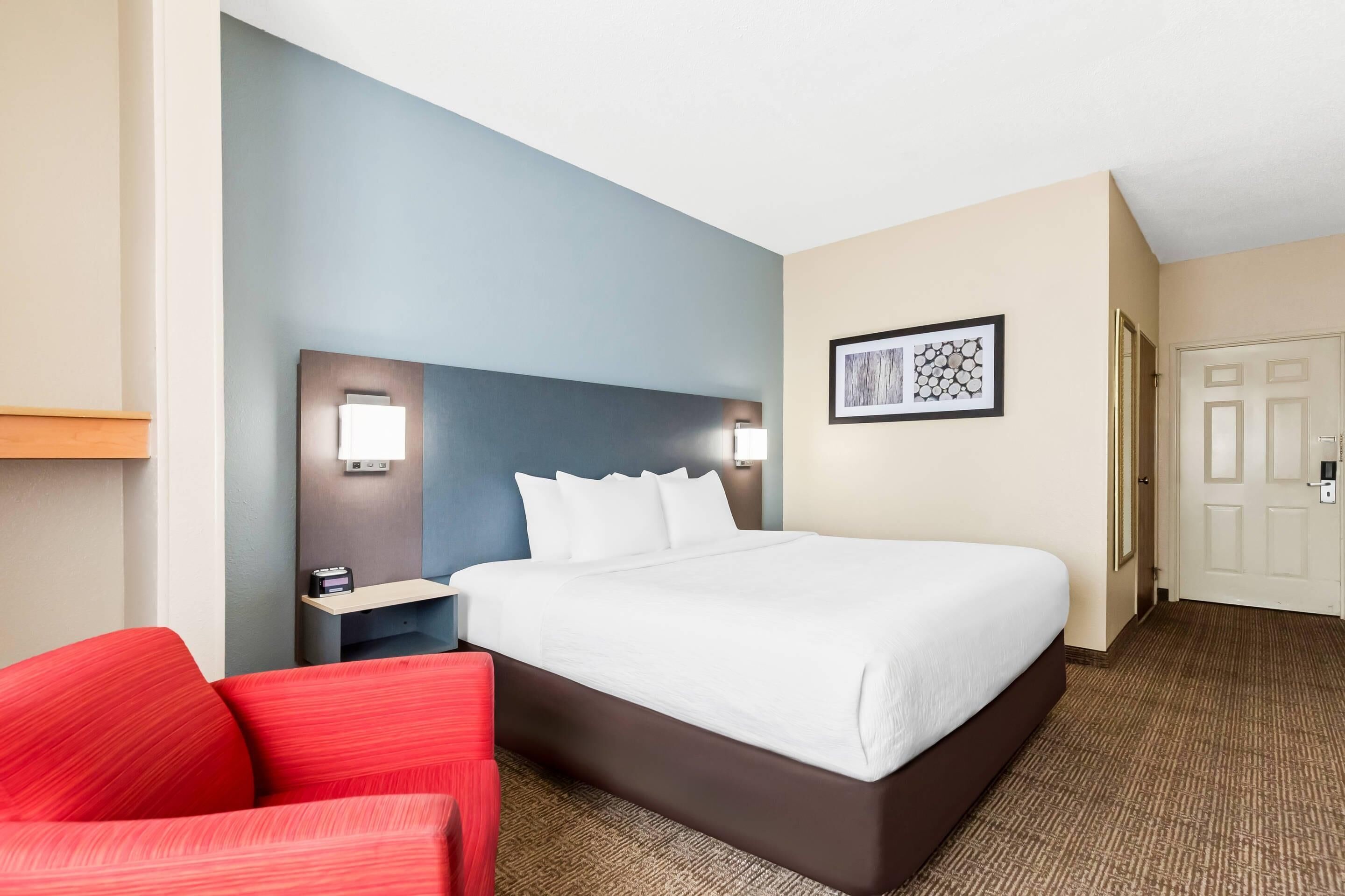 A hotel room with a large bed, a red chair, a nightstand with a lamp, and a framed picture on the wall.