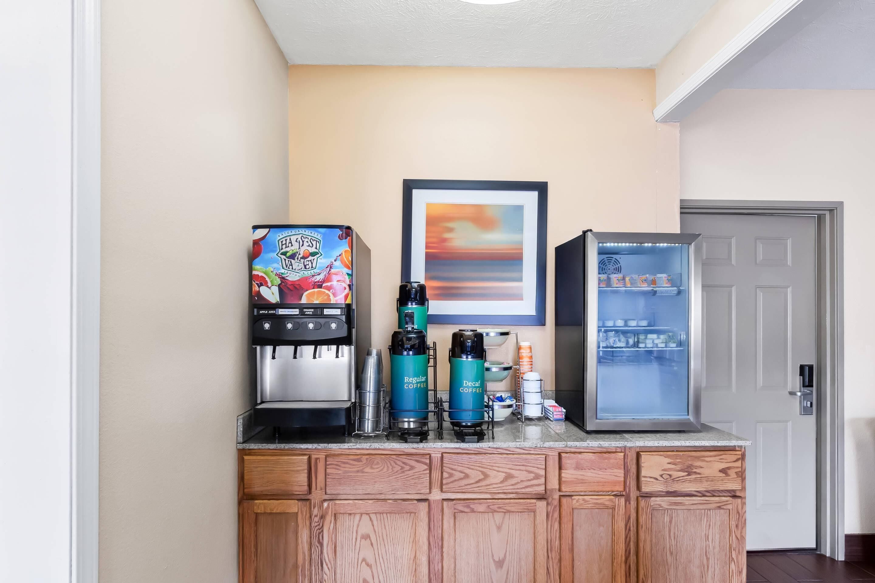 A kitchenette with a coffee maker, two coffee dispensers, a refrigerator, and a framed picture on the wall.