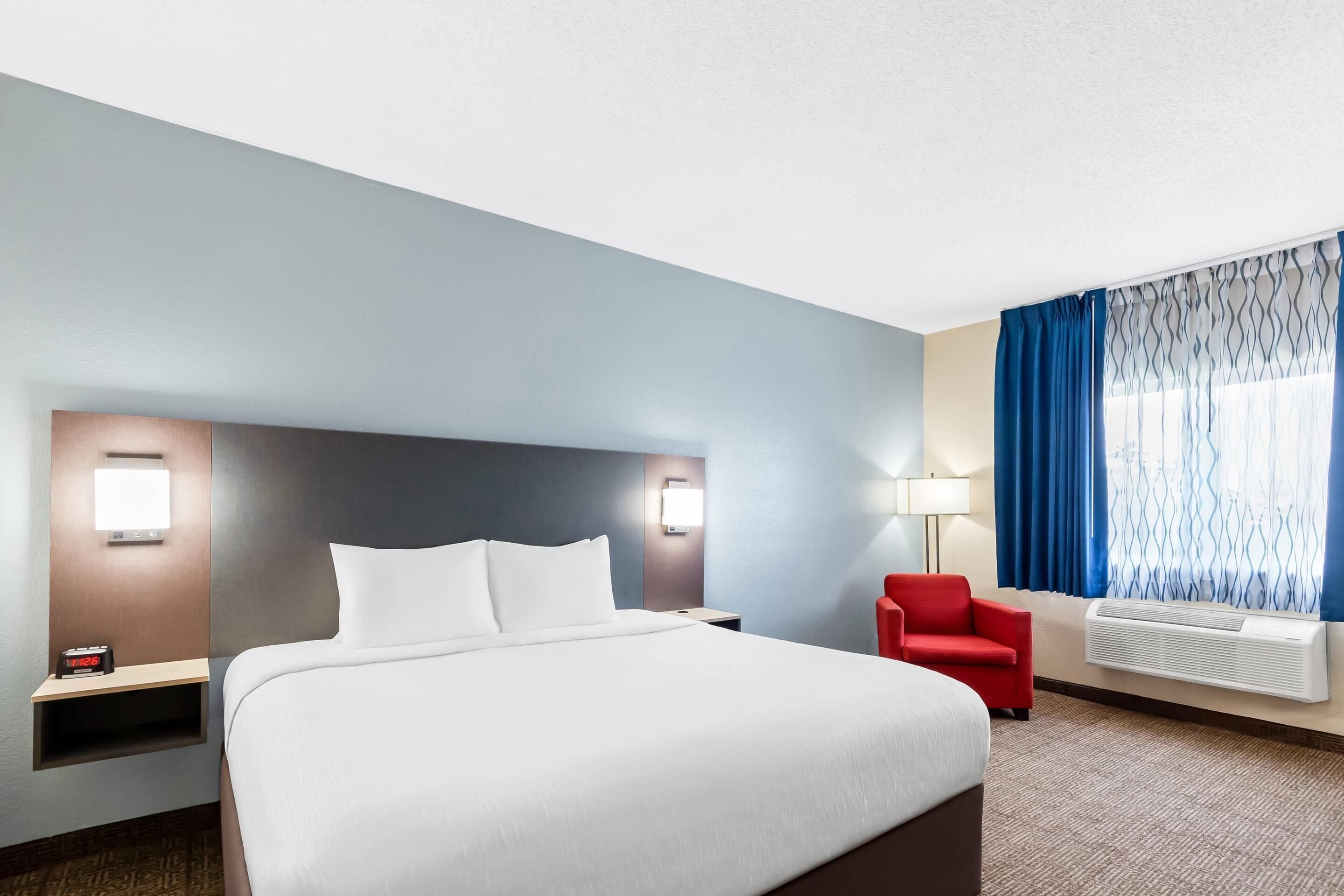 Una habitación de hotel con una cama grande, una silla roja, una mesita de noche con un reloj y una ventana con cortinas azules.
