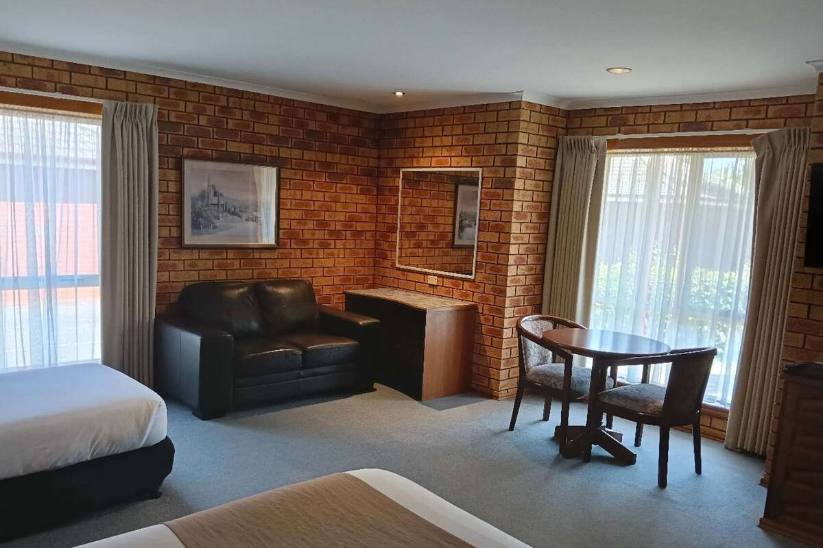 A hotel room with a brick wall, a bed, a sofa, a small table with chairs, and a window with curtains.