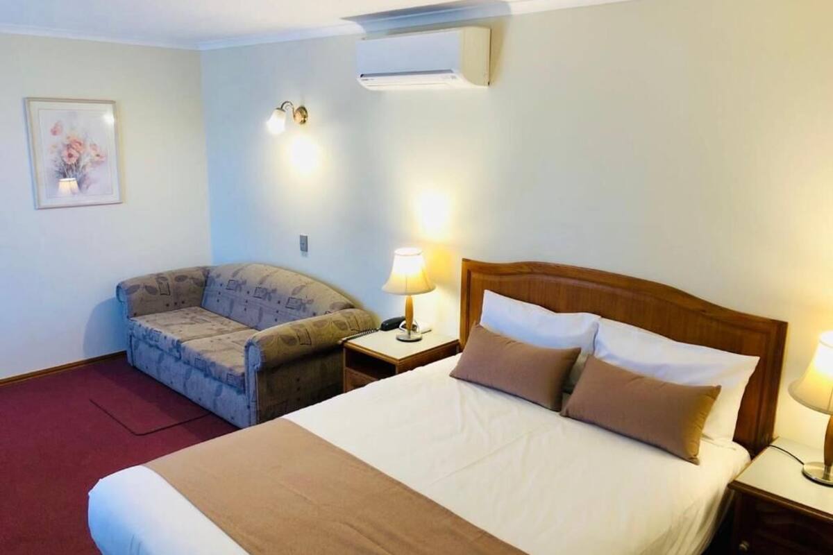 A hotel room with a large bed, a sofa, two bedside tables with lamps, an air conditioning unit, and a framed picture on the wall.