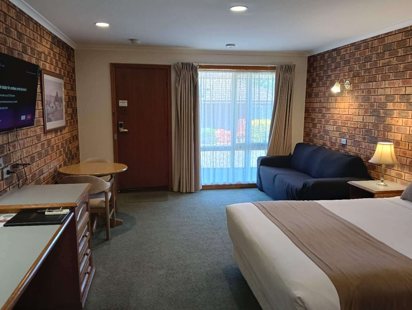 A hotel room with a bed, a desk, a chair, a sofa, a TV, and a window with curtains.