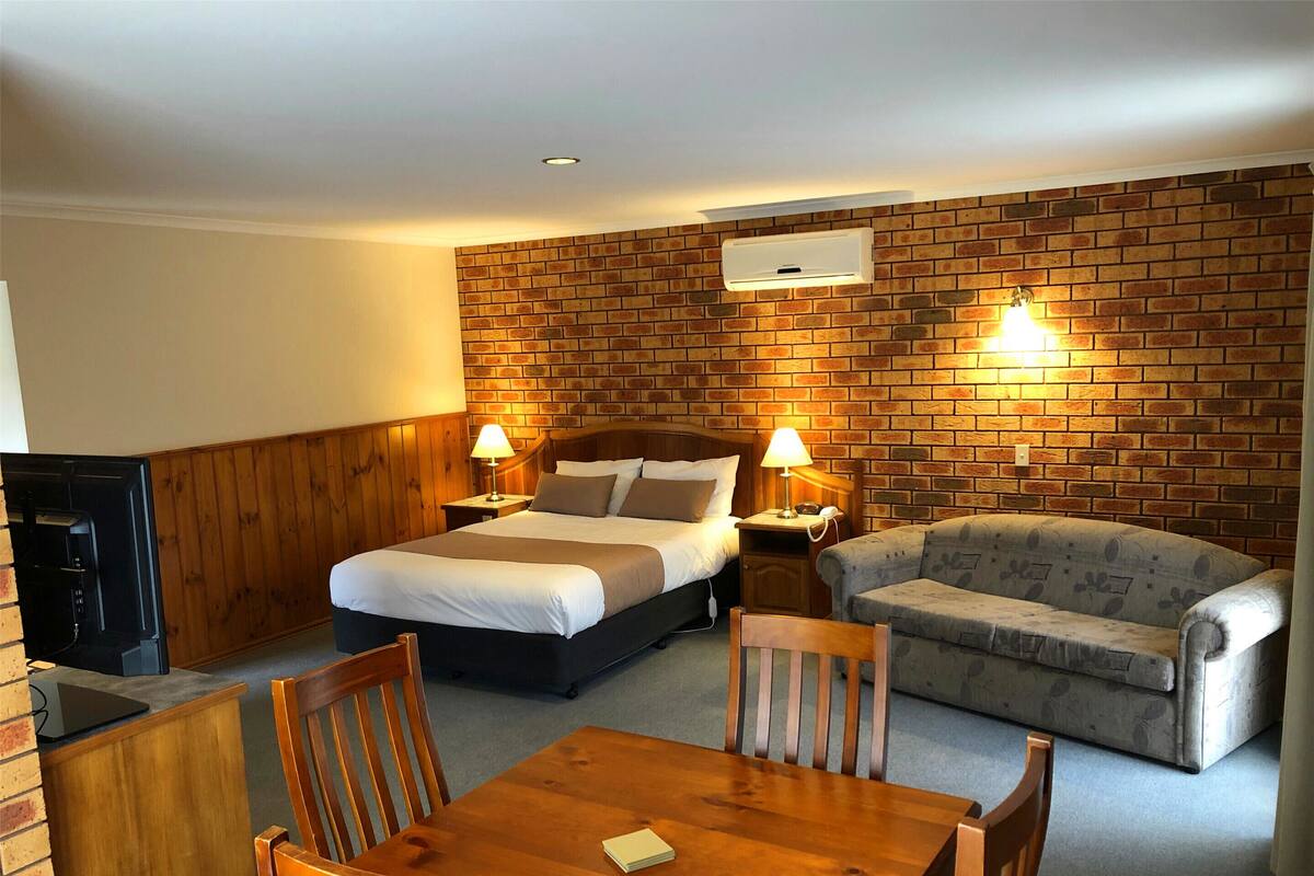 A hotel room with a brick wall, a bed, a sofa, a wooden table, and a TV.