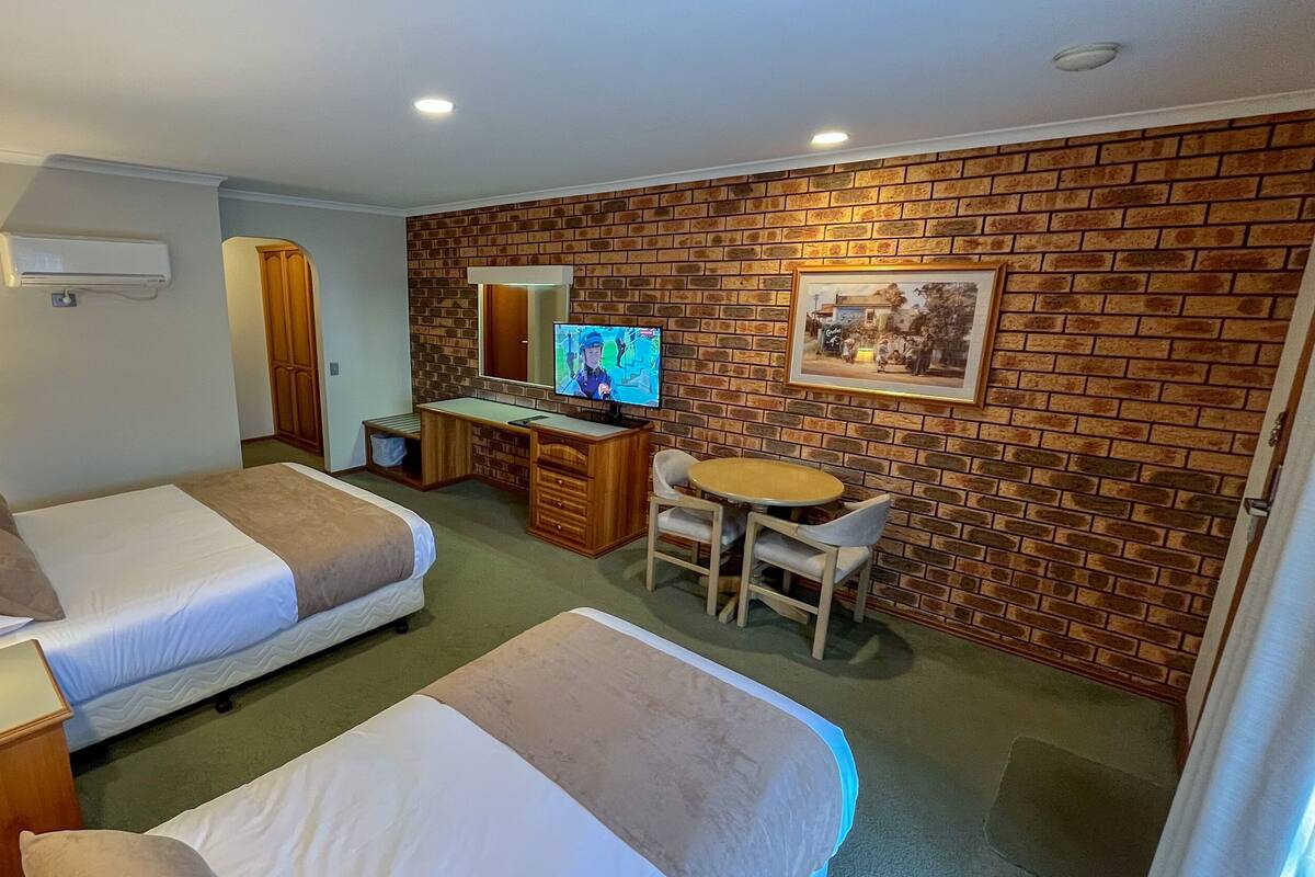 A hotel room with two beds, a desk with a computer, a chair, a TV, and a brick wall.