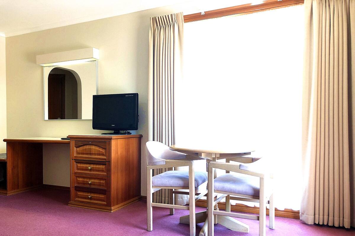 A hotel room with a wooden desk, a television, a chair, and a window with curtains.