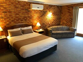 Habitación de hotel con pared de ladrillo, cama con ropa de cama blanca y marrón, una silla, una mesita de noche con lámpara y una ventana con cortinas.