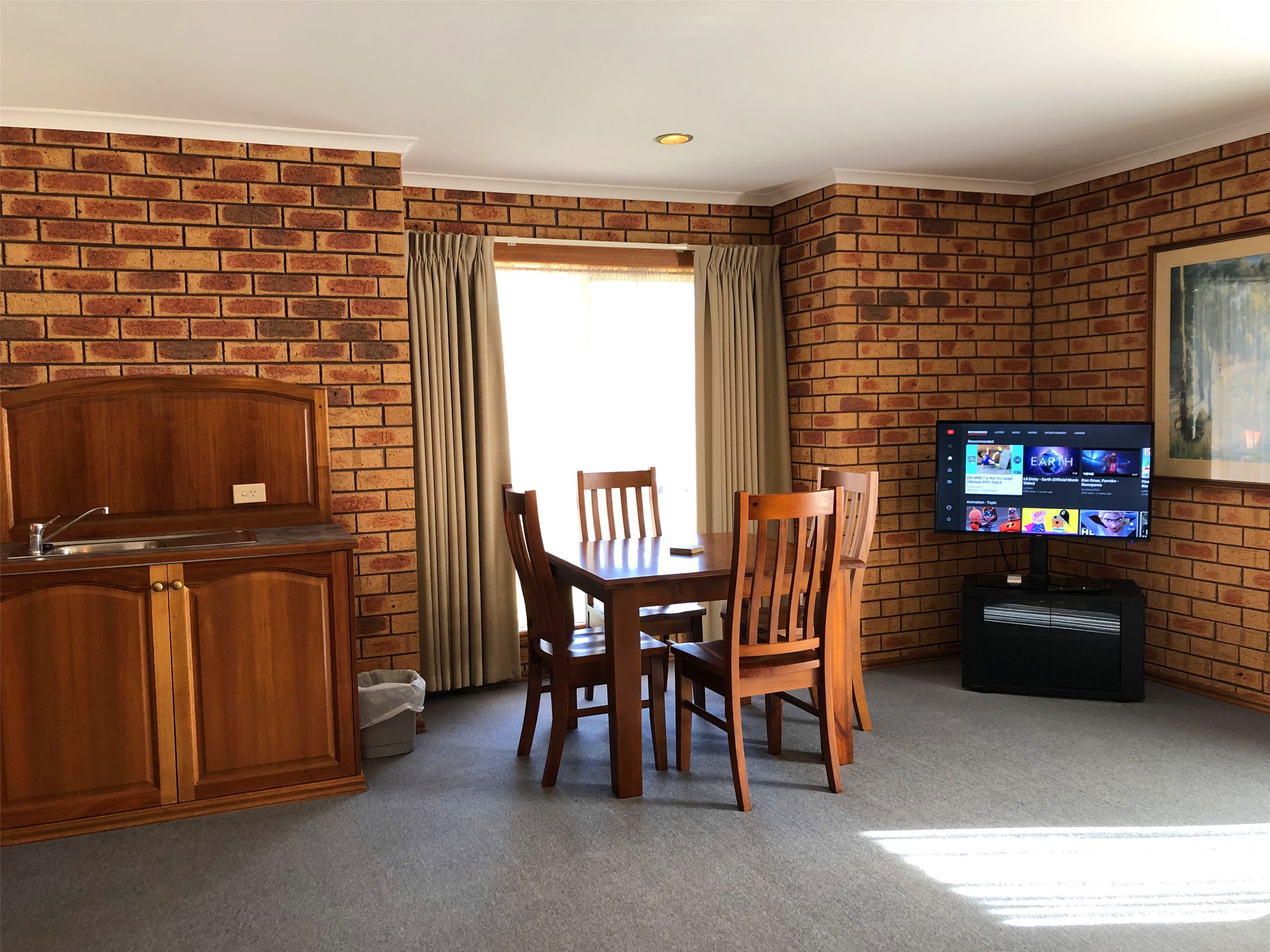 Una habitación con pared de ladrillo, una mesa de comedor de madera con sillas, un televisor sobre un mueble bajo y una ventana con cortinas.
