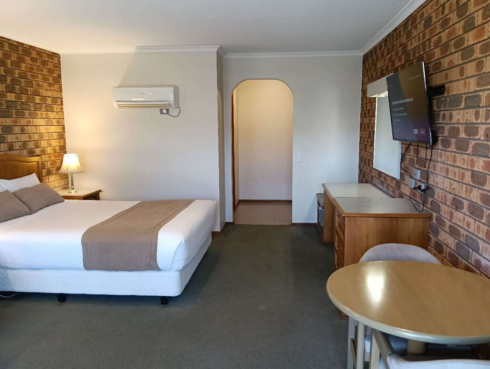 A hotel room with a bed, bedside table, lamp, desk, chair, TV, and brick wall.