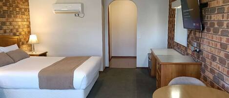 A hotel room with a bed, bedside table, lamp, desk, chair, TV, and brick wall.