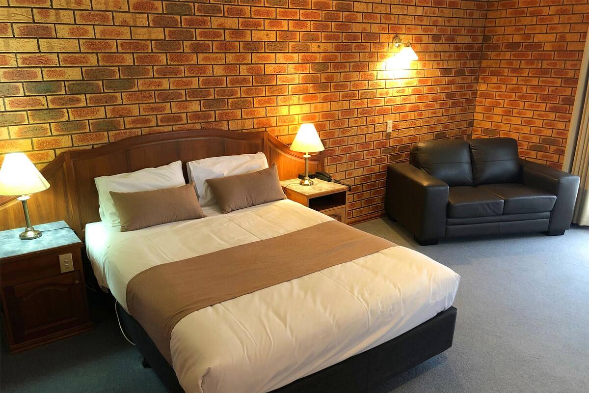 A hotel room with a brick wall, a bed with white and brown bedding, two bedside lamps, a wooden nightstand, a black leather armchair, and a small desk with a lamp.