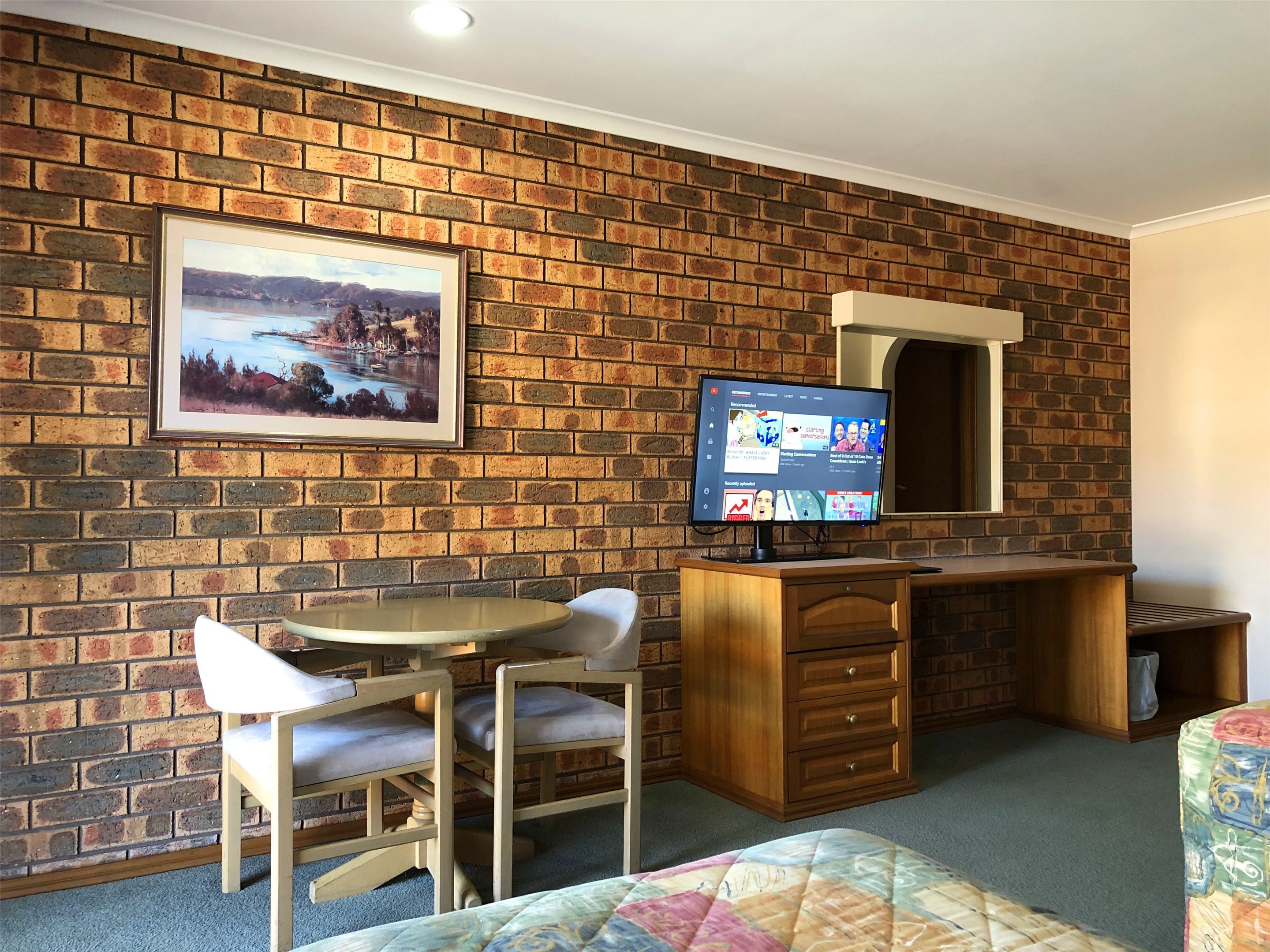 A room with a brick wall, a framed landscape painting, a wooden desk with a computer monitor, a chair, and a bed with a colorful quilt.