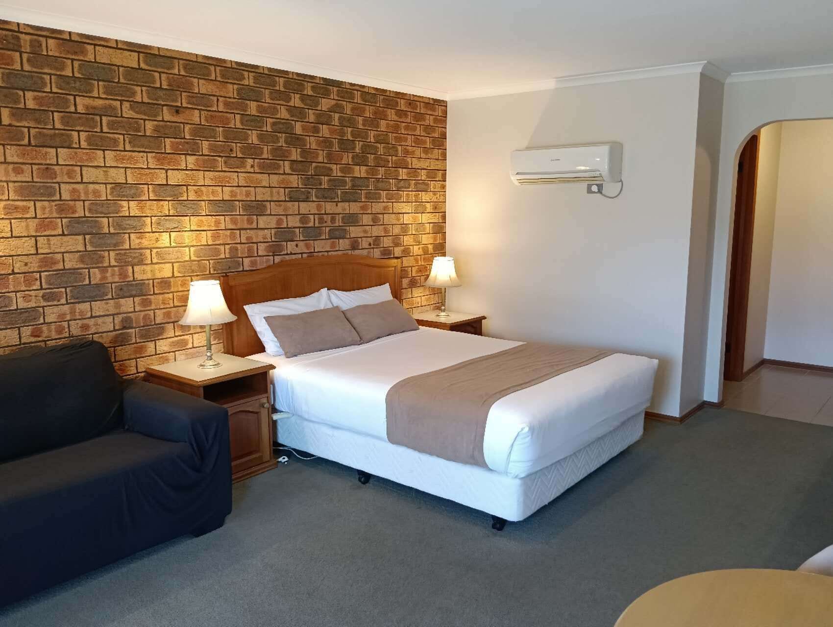 A hotel room with a brick wall, a bed with white bedding, two bedside lamps, a dark sofa, and a wall-mounted air conditioner.