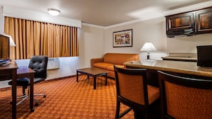A hotel room with a seating area, a small table, a desk with a chair, a television, a window with curtains, a framed picture on the wall, and a kitchenette with a microwave.