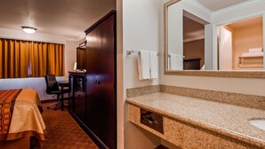 A hotel room with a bed, a desk, a chair, a wardrobe, and a bathroom with a sink and mirror.