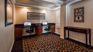 A hotel room with a desk, two chairs, a laptop, a desktop computer, a decorative wall piece, and a patterned carpet.