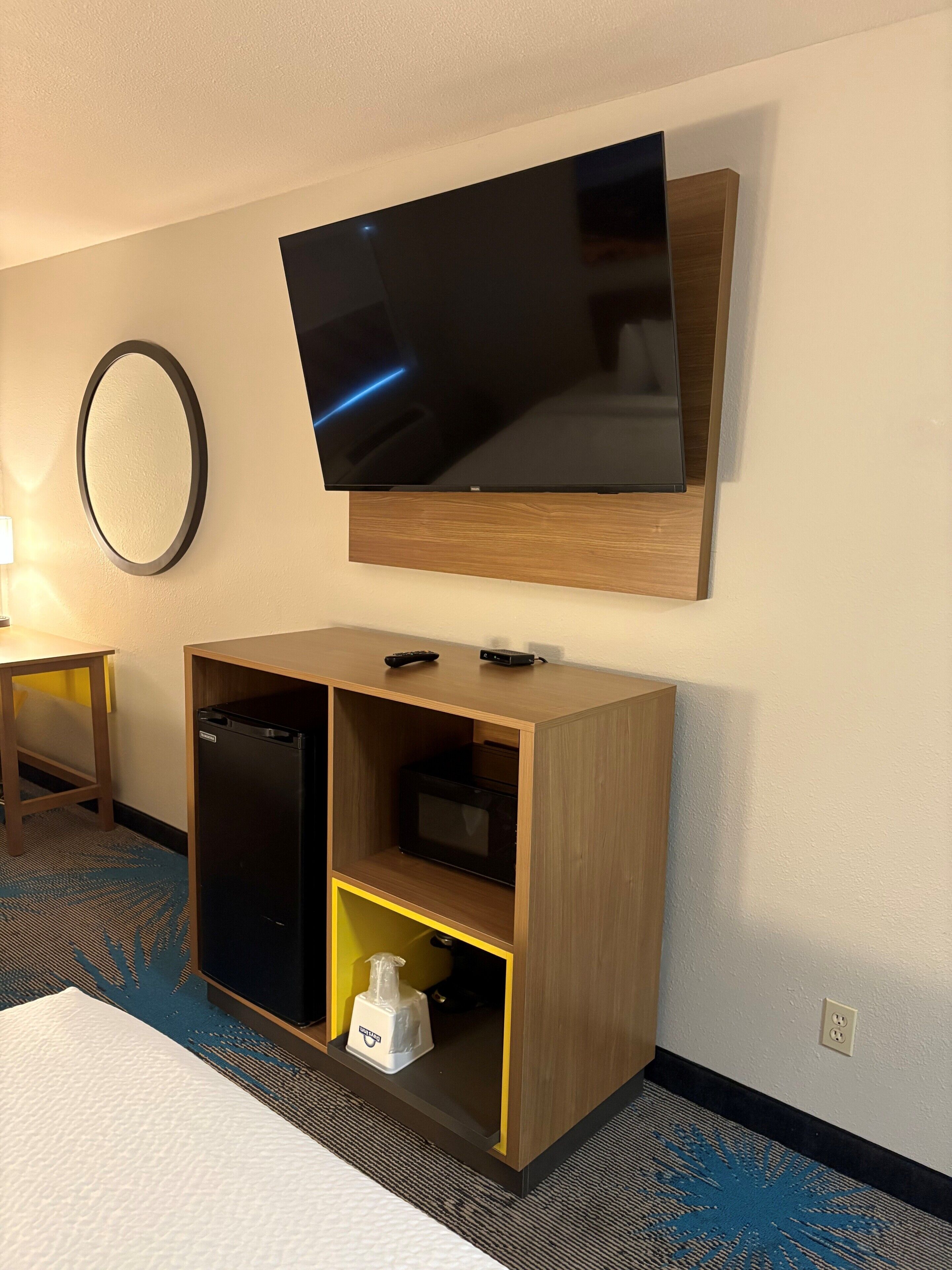 Quarto de hotel com televisão de tela plana na parede, um armário de madeira com micro-ondas e uma pequena mesa.