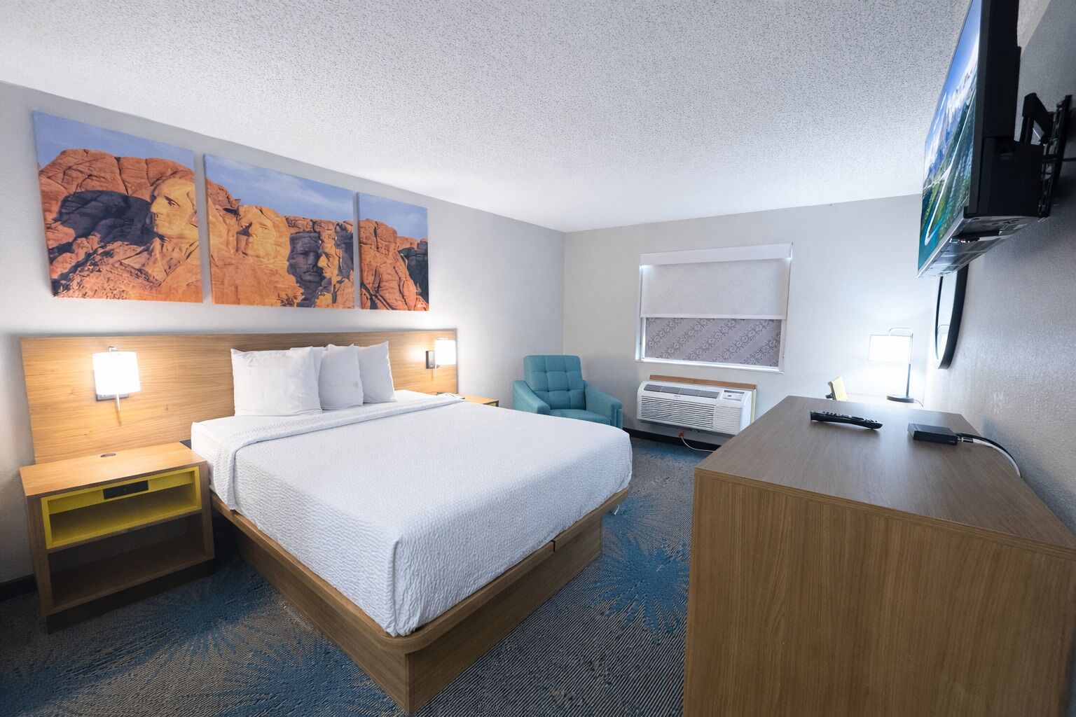 A hotel room with a bed, a desk, a chair, an air conditioning unit, and artwork on the wall.