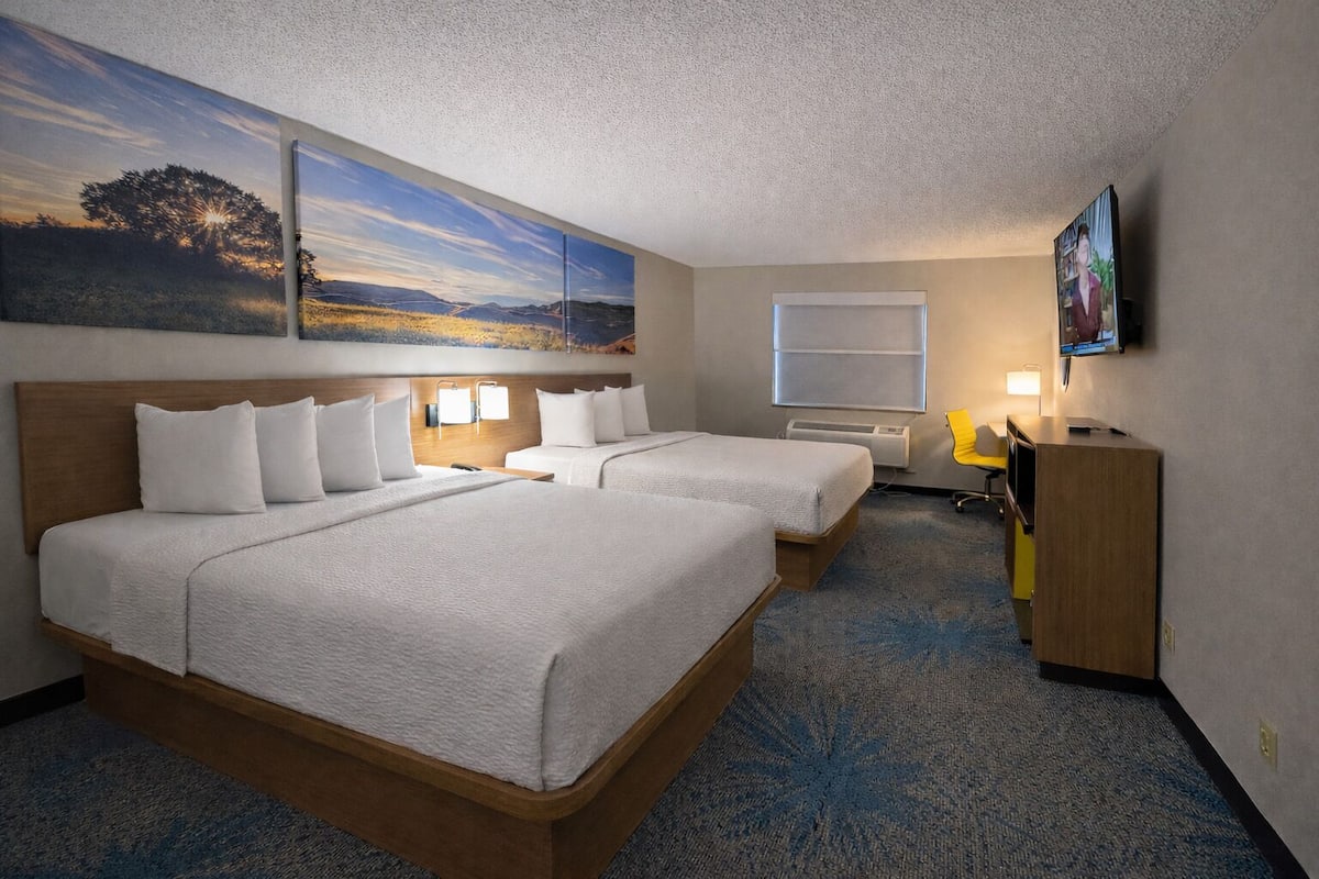 A hotel room with two beds, a desk, a chair, a TV, and artwork on the walls.