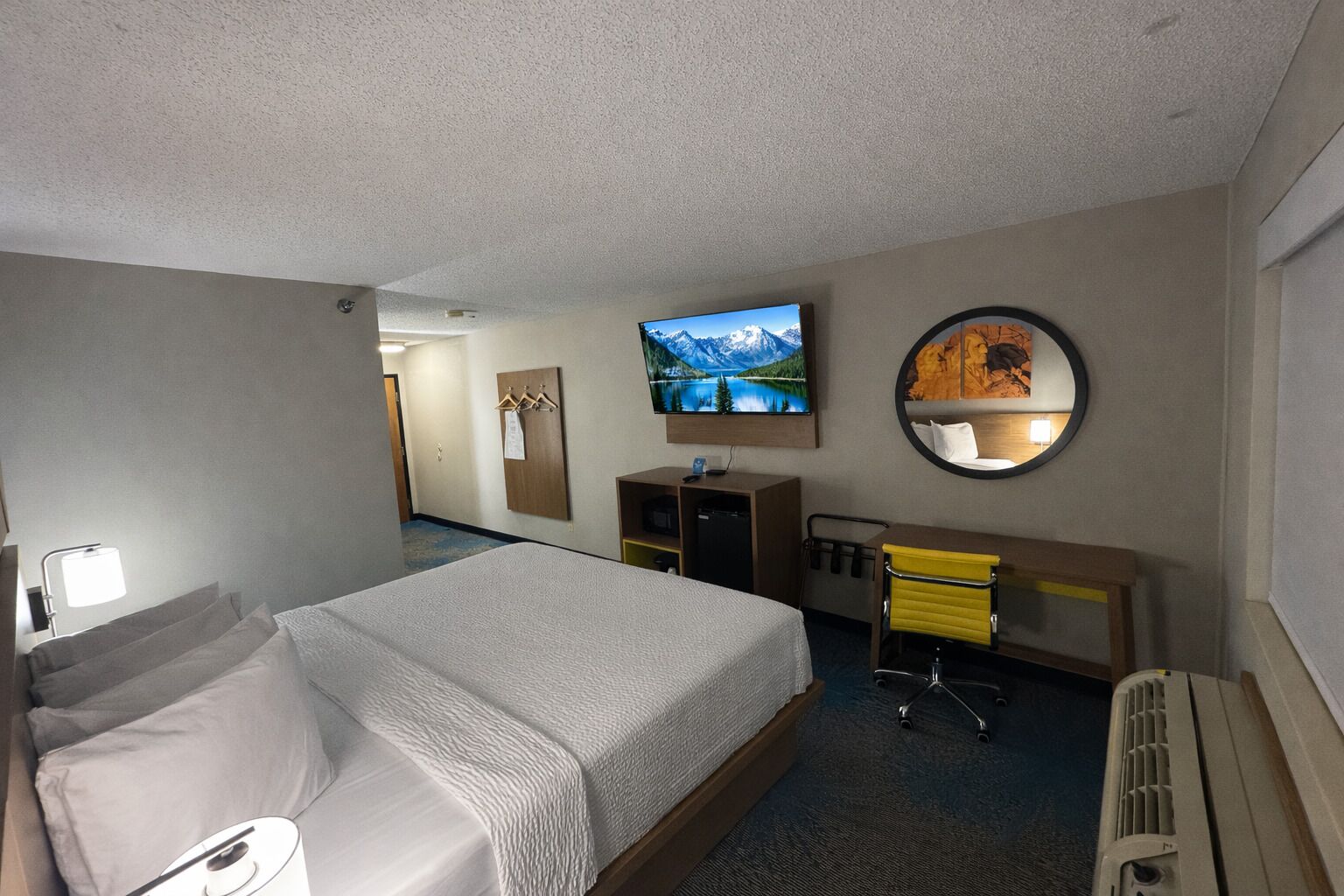 Une chambre d’hôtel avec un grand lit, un bureau avec une chaise, une télévision, un miroir et un porte-manteau.