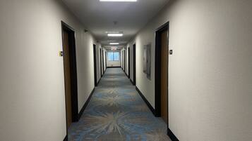 Corredor longo e estreito de hotel com carpete estampado, portas em ambos os lados e uma claraboia no final.