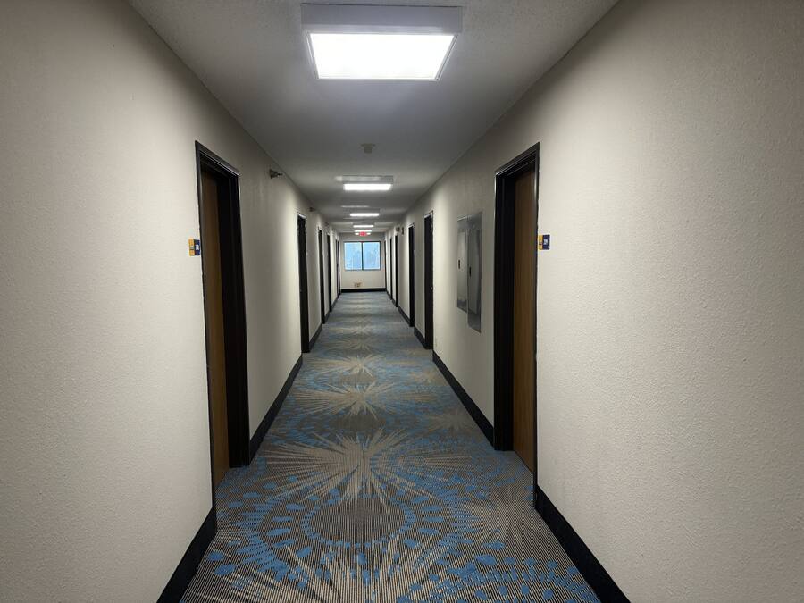 Corredor longo e estreito de hotel com carpete estampado, portas em ambos os lados e uma claraboia no final.