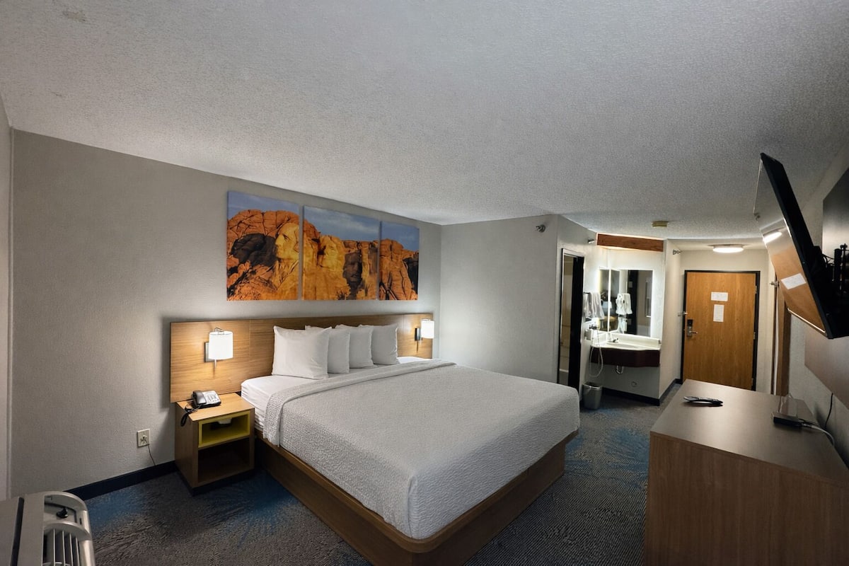 A hotel room with a large bed, bedside lamps, a desk, a mirror, and a bathroom visible through an open door.
