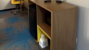 A hotel room with a TV mounted on the wall, a desk with a chair, a lamp, and a minibar.