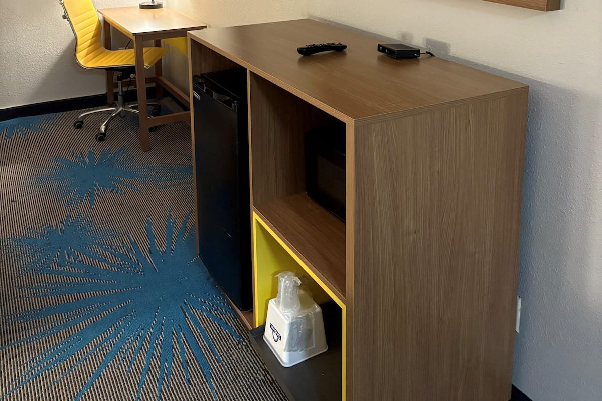 A hotel room with a TV mounted on the wall, a desk with a chair, a lamp, and a minibar.