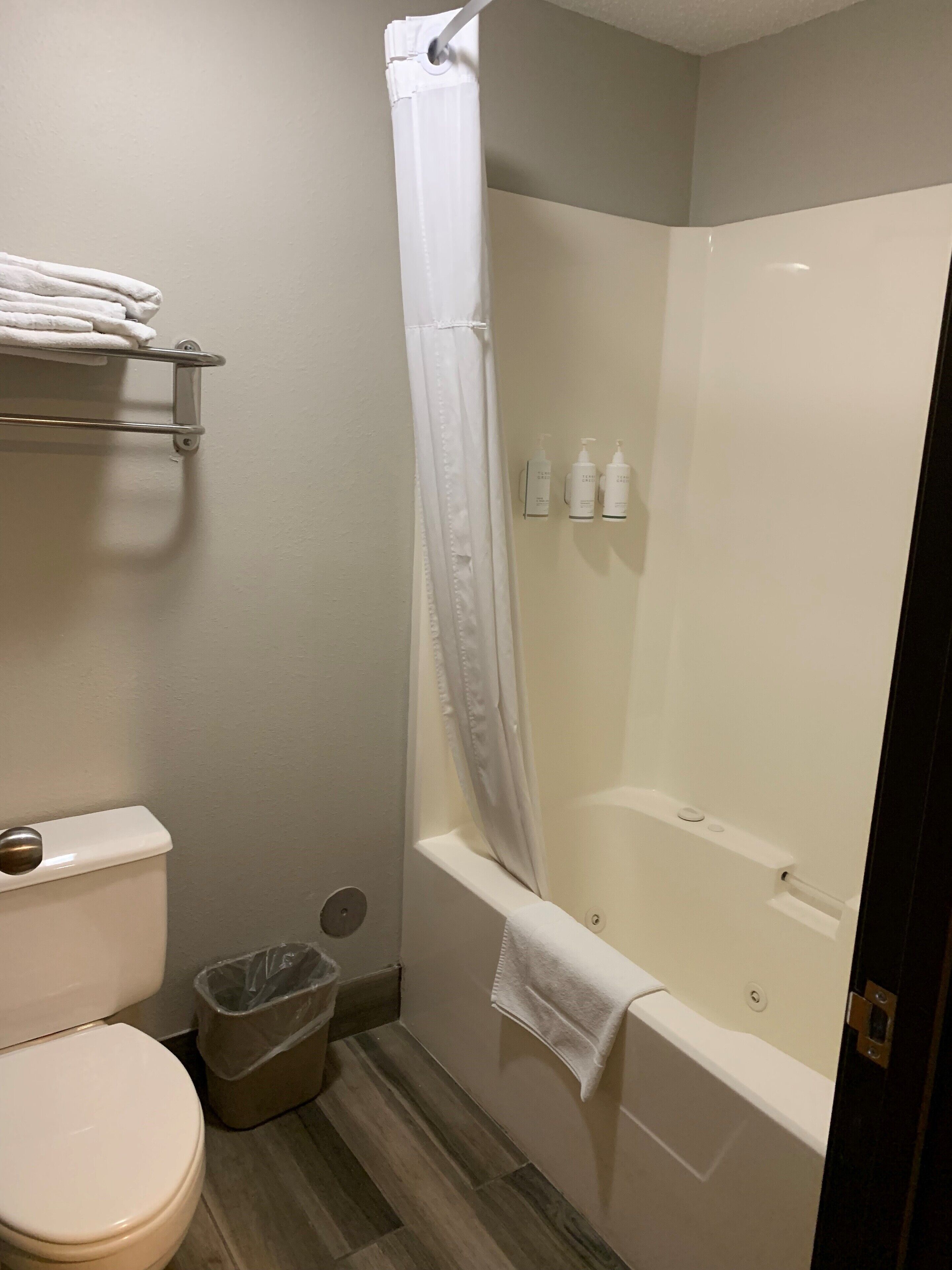 Een badkamer met een toilet, een douche met een witte gordijn, een plank met handdoeken en een prullenbak.