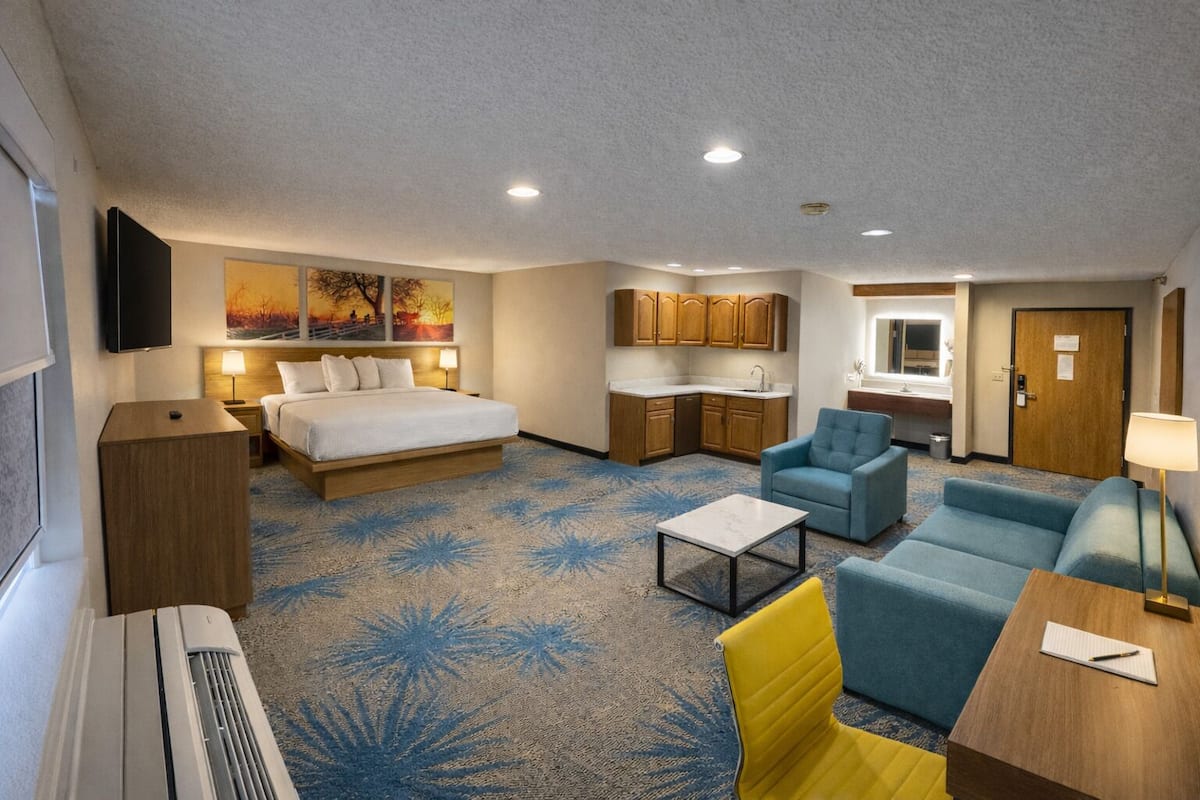 A hotel room with a bed, a sofa, a chair, a small table, a desk, a kitchenette, and a bathroom.