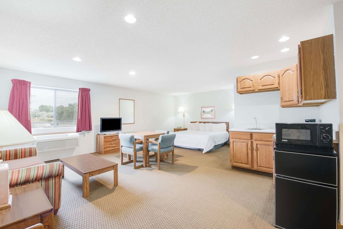 A hotel room with a bed, a TV, a dining table with chairs, a kitchen area with a microwave and stove, and a window with curtains.