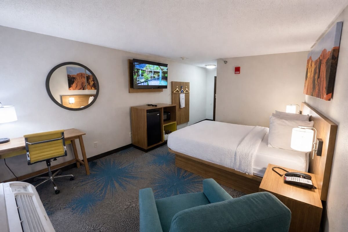 A hotel room with a bed, a desk with a chair, a television, a wardrobe, and a small table with a lamp.