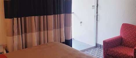 A hotel room with a bed, curtains, a red chair, and a view of the outside.