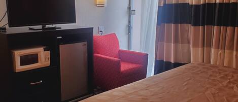 A hotel room with a bed, a TV, a microwave, a red chair, and a window with curtains.