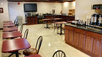 A restaurant with multiple tables and chairs, a kitchen area with a coffee machine, and a television mounted on the wall.