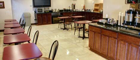 A restaurant with multiple tables and chairs, a kitchen area with a coffee machine, and a television mounted on the wall.