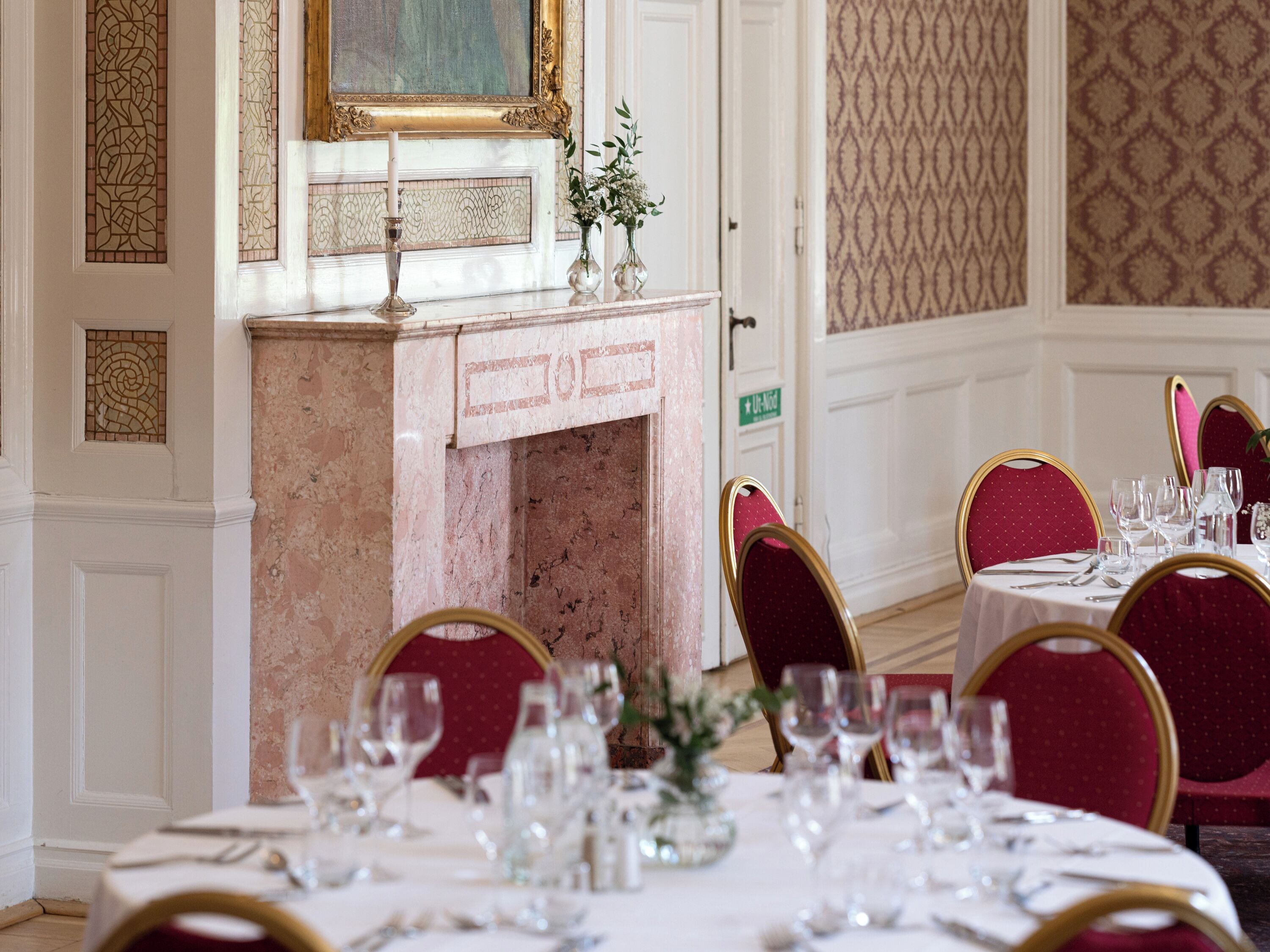 A dining area with marble fireplace, red upholstered chairs, and a white table set with wine glasses and cutlery.