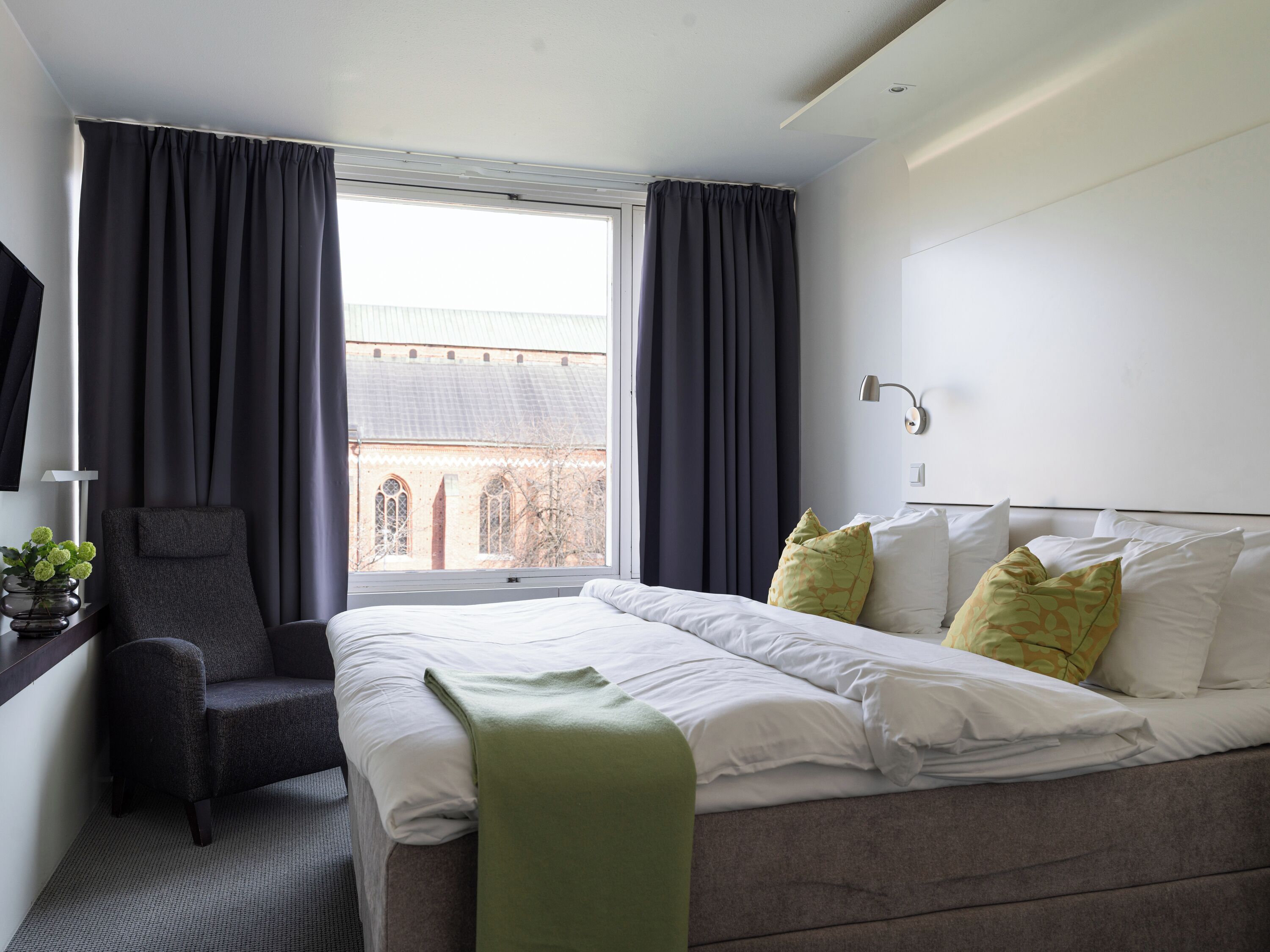 A modern hotel room with a large bed, a desk with a lamp, a chair, and a view of a building through the window.