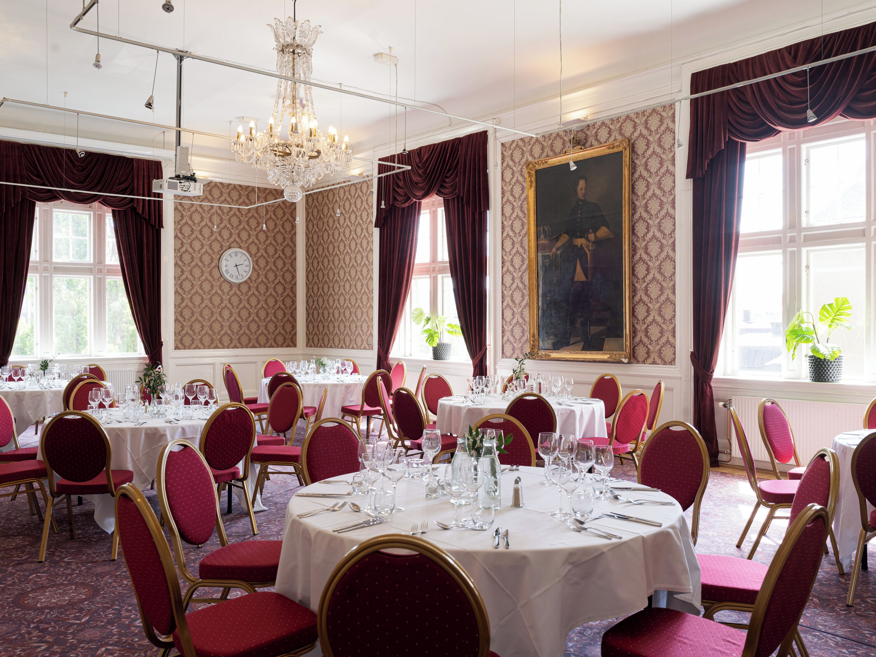 A formal dining room with multiple round tables set for service, red upholstered chairs, and a large chandelier.