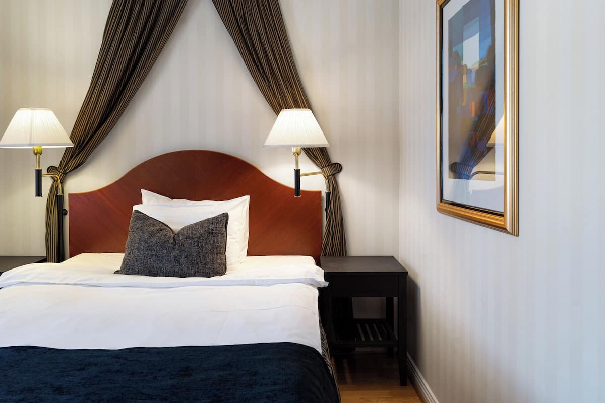 Ein Hotelzimmer mit einem großen Bett, zwei Nachttischlampen, einem dunklen Holz-Kopfteil, einem kleinen Beistelltisch und einem gerahmten Bild an der Wand.
