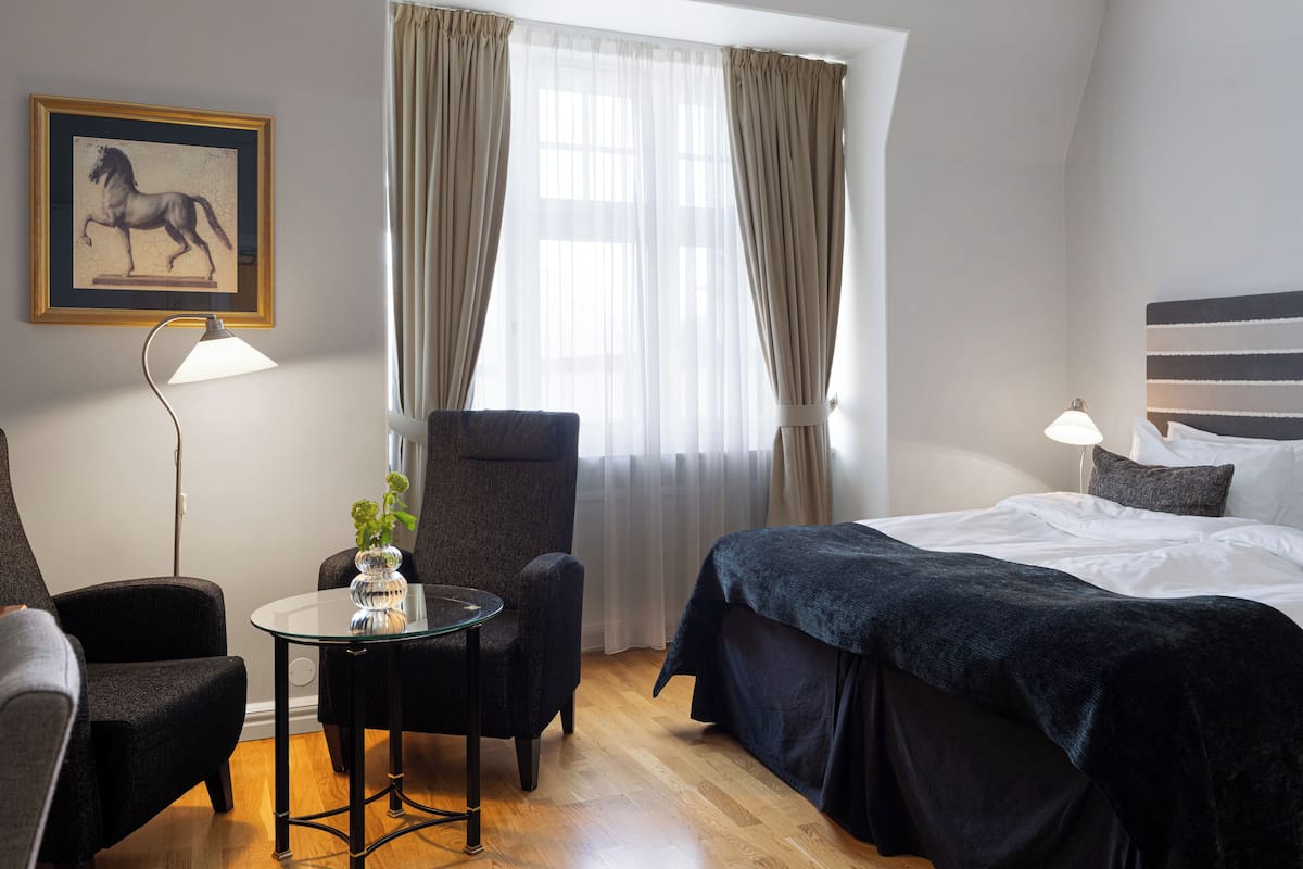 Ein Hotelzimmer mit einem Bett, zwei Sesseln, einem kleinen Tisch mit einer Vase, einer Lampe und einem Fenster mit Vorhängen.