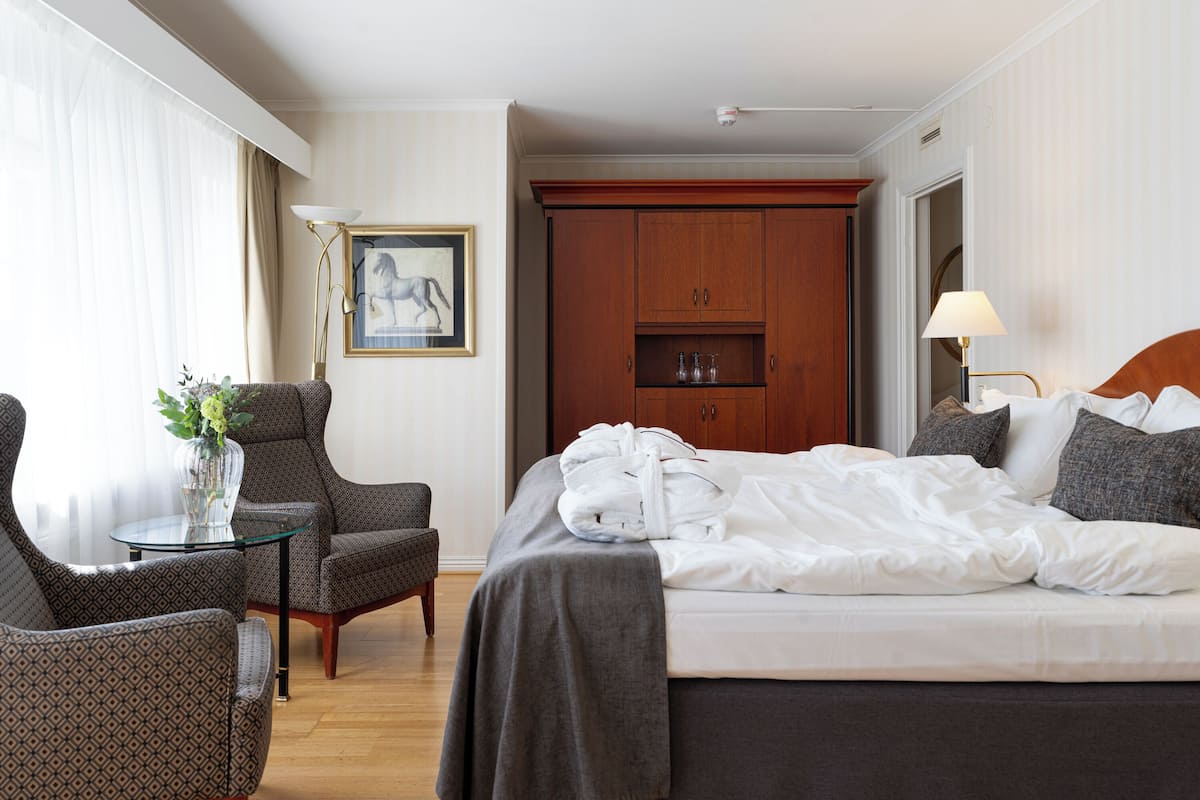 Ein Hotelzimmer mit Bett, einem Sessel, einem Tisch mit Vase sowie einem Holzschrank und einem gerahmten Bild an der Wand.