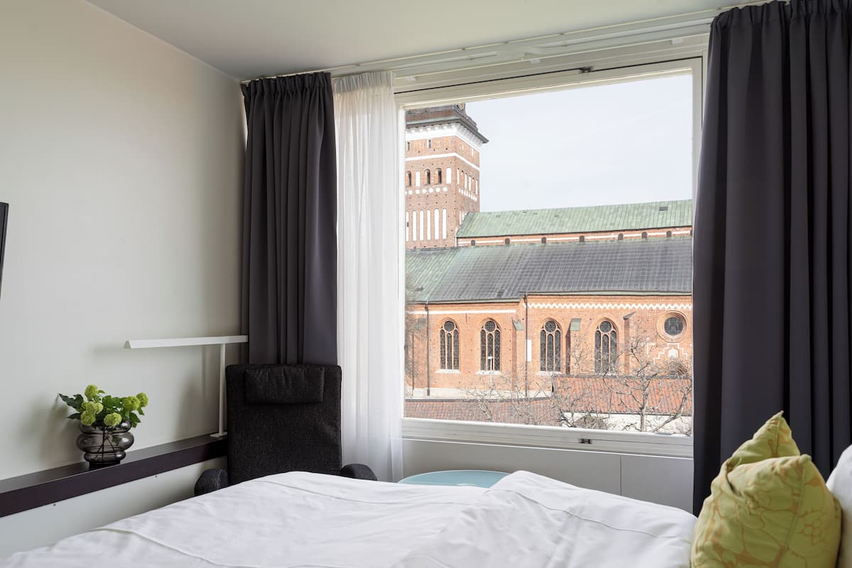 Ein Hotelzimmer mit Bett, Sessel, kleinem Tisch und Blick auf ein Backsteingebäude durch das Fenster.