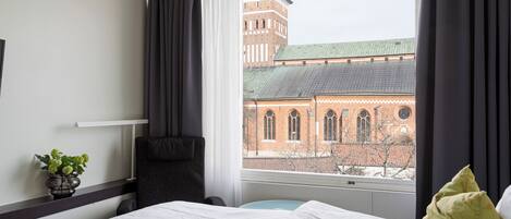 A hotel room with a bed, a chair, a small table, and a view of a brick building through the window.