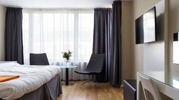 A hotel room with a bed, a desk, a chair, a small table with a vase, and a TV on the wall.