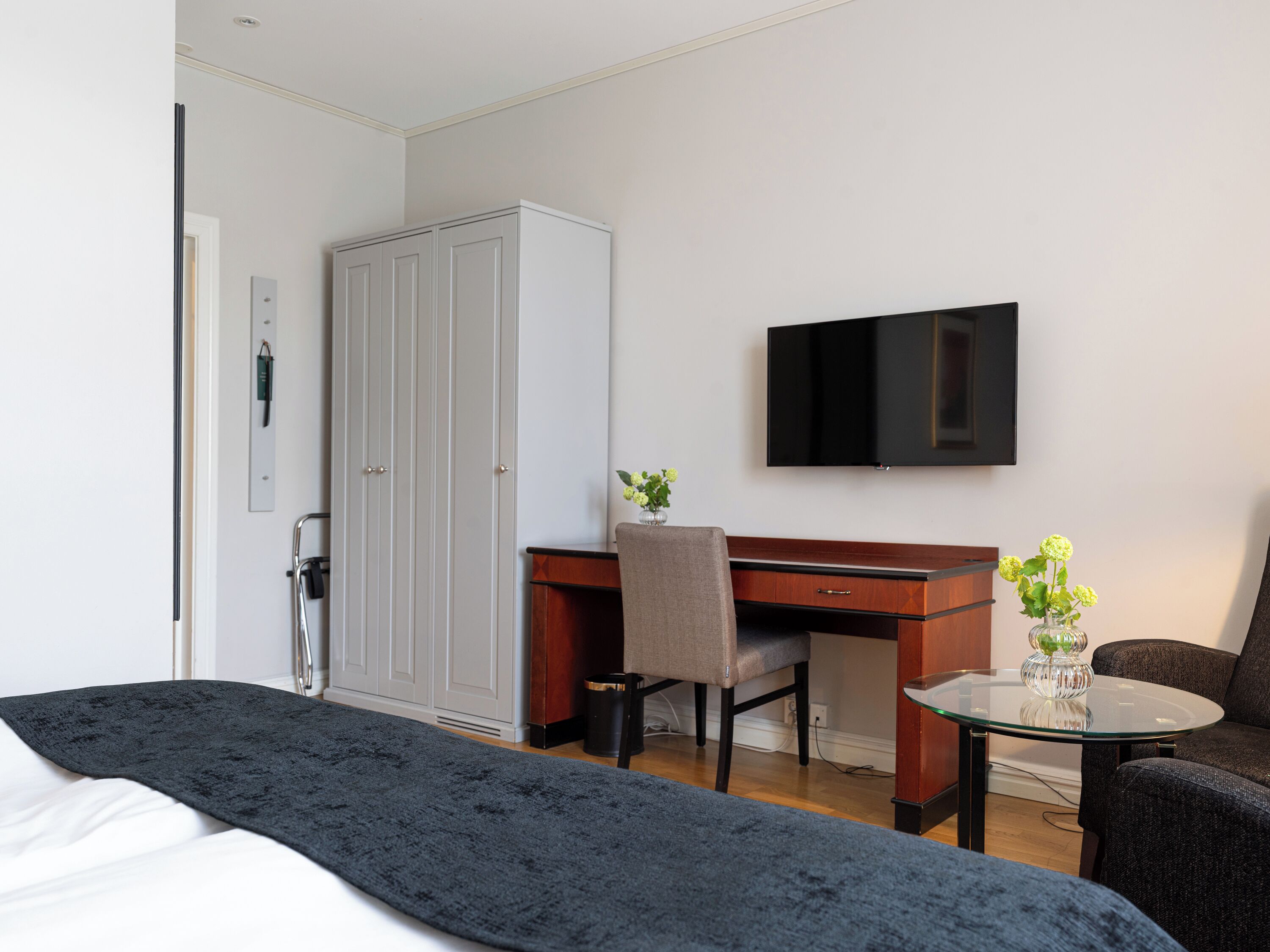 A hotel room with a bed, a desk, a chair, a TV, and a small table with a vase of flowers.