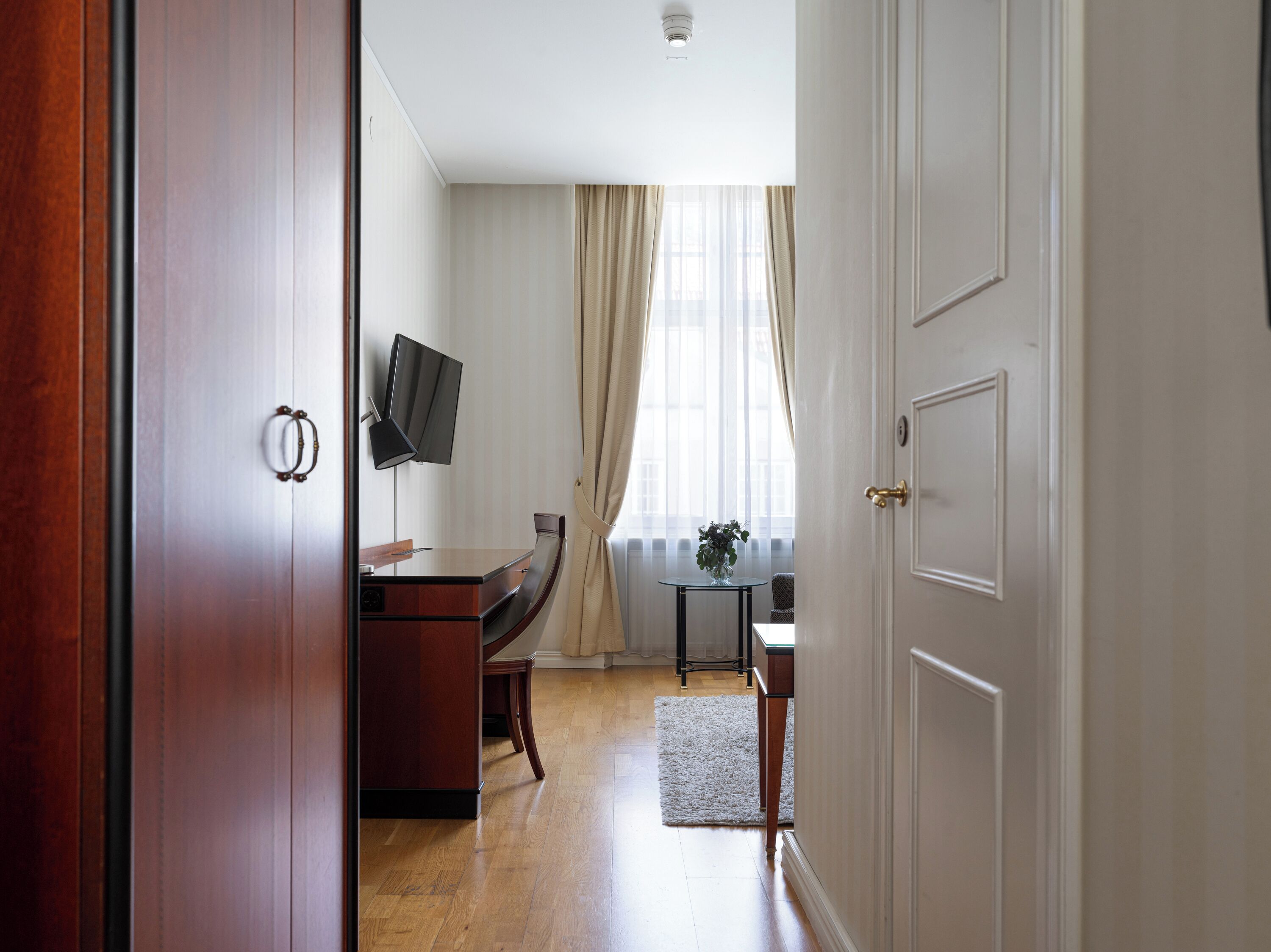 Habitación de hotel con escritorio de madera, televisor montado en la pared, una mesita con un florero y una ventana con cortinas.