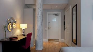 Habitación de hotel con un pilar de mármol, un escritorio con lámpara, una silla roja, una cama y una puerta que da a otra habitación.