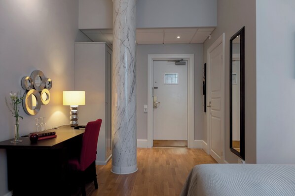 A hotel room with a marble column, a desk with a lamp, a red chair, a bed, and a door leading to another room.
