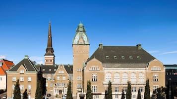 A historic building with a prominent tower and a large central structure, surrounded by parked cars and trees.